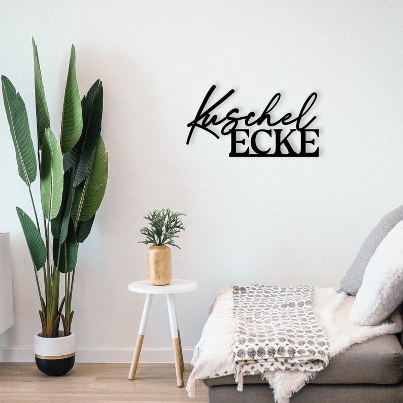 May include: Black metal wall decor featuring the words "Kuschel Ecke" in a cursive font. The decor is mounted on a white wall above a white sofa with a black and white patterned throw.