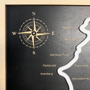 May include: A black wooden map with a white outline of a region. The map includes the names of places like Adenauer Forst, Fuchsroh, and Schwedenkreuz. A compass rose is in the top left corner with the directions North, South, East, and West.