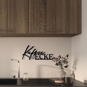 May include: Black metal wall decor with the text "Kaffee Ecke" in a stylised font. The decor is mounted on a white wall above a kitchen sink.