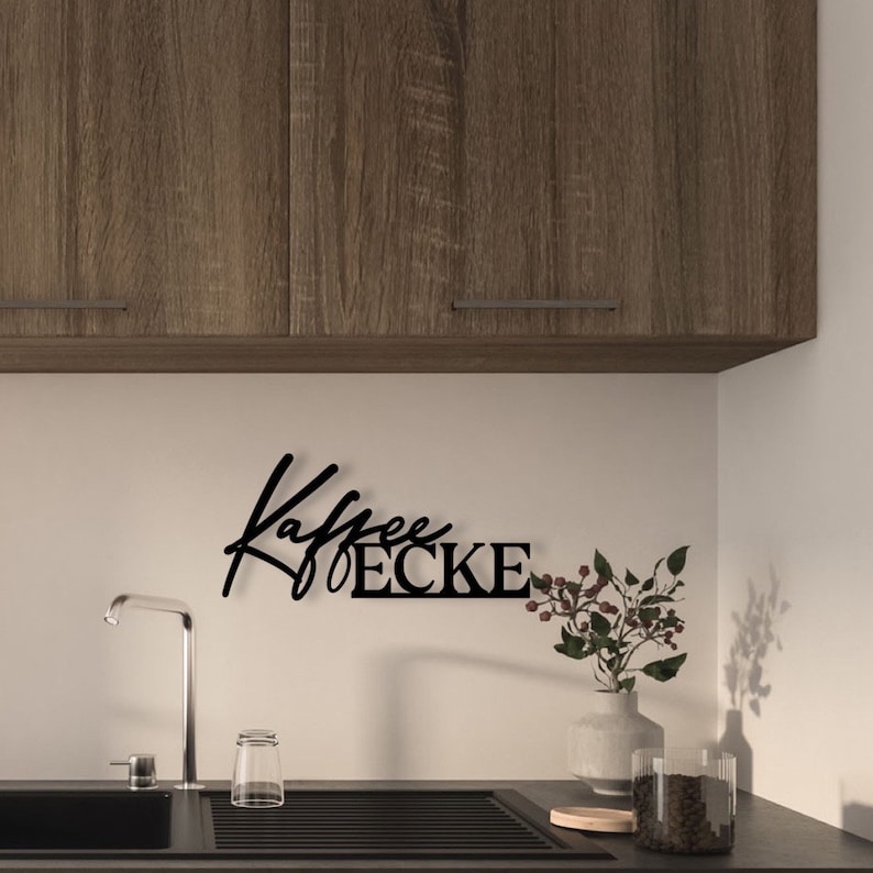 May include: Black metal wall decor with the text "Kaffee Ecke" in a stylised font. The decor is mounted on a white wall above a kitchen sink.