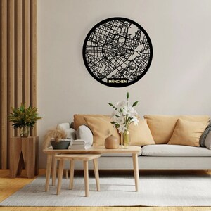 May include: Black and white map of Munich, Germany, in a circular metal frame, hanging on a white wall above a light gray couch with tan pillows. The couch is in a living room setting with a wooden coffee table and two side tables.