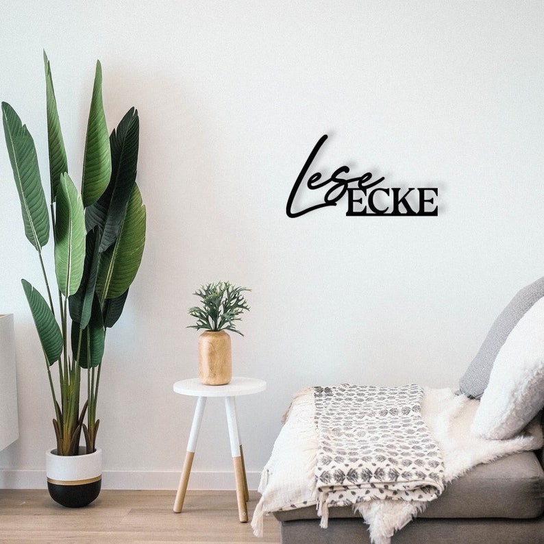 May include: Black metal wall decor featuring the text "Leser Ecke" in a contemporary font style. The decor is mounted on a white wall above a white sofa with a grey throw blanket.