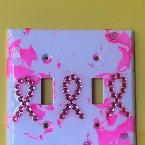 May include: A decorative light switch cover with a white base, decorated with pink paint splatters and three pink rhinestone ribbons. Small pink hearts with blue outlines are scattered. It has two switch openings.
