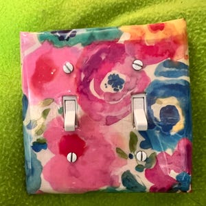 May include: A decorative light switch cover featuring a watercolor floral design. The cover has a glossy finish and showcases pink, blue, and purple flowers with green leaves. Two white toggle switches are visible, secured by silver screws.