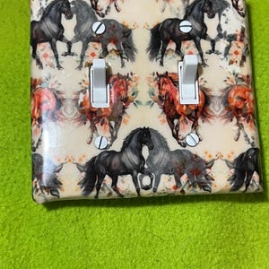 May include: A decorative light switch cover featuring a repeating pattern of horses in various colors, including black, gray, and brown, set against a cream background with floral accents. The cover has two toggle switches and is mounted with silver screws.