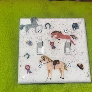 May include: White light switch cover decorated with horse-themed embellishments. Features three horse figurines, horseshoes, ribbons, and a riding helmet. The cover has two light switches and is covered in glitter.