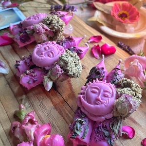 May include: Pink and purple candles with dried flowers and herbs. The candles are shaped like a crescent moon with a face on it.