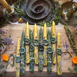 May include: A collection of eight taper candles in shades of yellow and forest green, each decorated with embedded dried flowers, small crystals, and a prominent dark green wax spiral accent. The candles are arranged vertically on a wooden surface.