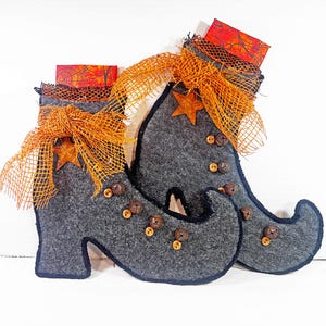 May include: Gray felt witch boots with orange accents, perfect for Halloween decor. The boots feature orange mesh ribbon, star-shaped embellishments, and small brown bells. The tops of the boots have orange and black patterned fabric.