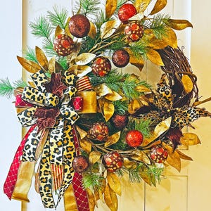 May include: A gold and brown Christmas wreath with a leopard print bow. The wreath is decorated with gold leaves, red and brown ornaments, and a brown velvet flower.