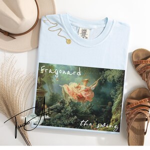 May include: Light blue t-shirt featuring the painting "The Swing" by Fragonard. The shirt has a crew neck and short sleeves. A gold necklace and brown sandals are also in the image, along with a beige hat and decorative grass.
