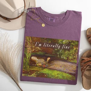 May include: A purple t-shirt with the text "I'm literally fine" over a painting of a person in water. The image also includes a beige hat, a gold necklace, brown sandals, and decorative dried grass.