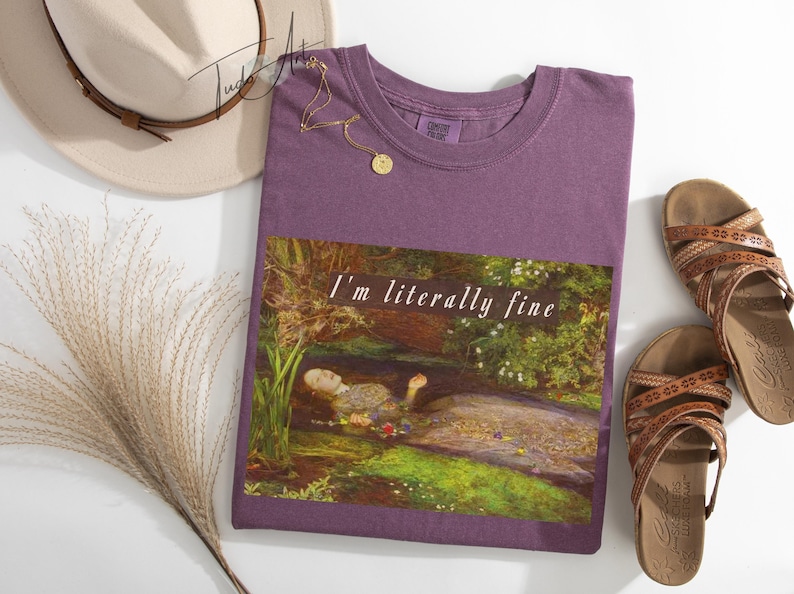 May include: A purple t-shirt with the text "I'm literally fine" over a painting of a person in water. The image also includes a beige hat, a gold necklace, brown sandals, and decorative dried grass.