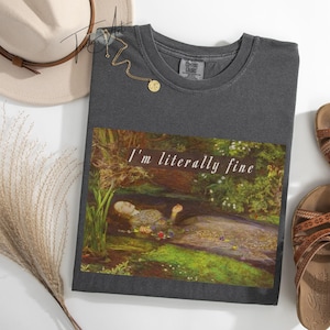 May include: Dark grey t-shirt with the text "I'm literally fine" above a painting of a woman in a stream. The image includes a hat, sandals, and a necklace.