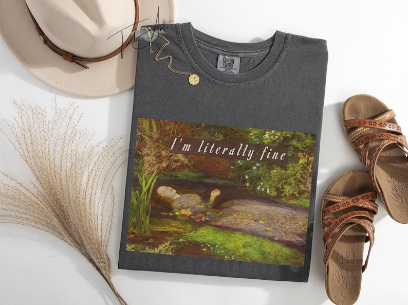 May include: Dark grey t-shirt with the text "I'm literally fine" above a painting of a woman in a stream. The image includes a hat, sandals, and a necklace.