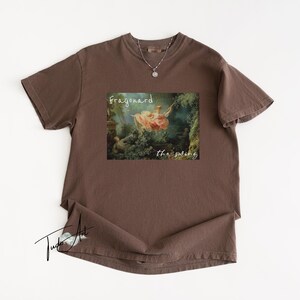 May include: A brown t-shirt featuring a print of a classical painting with the words "Fragonard" and "the swing". A silver necklace with a pendant is visible. The shirt is laid flat on a white surface.