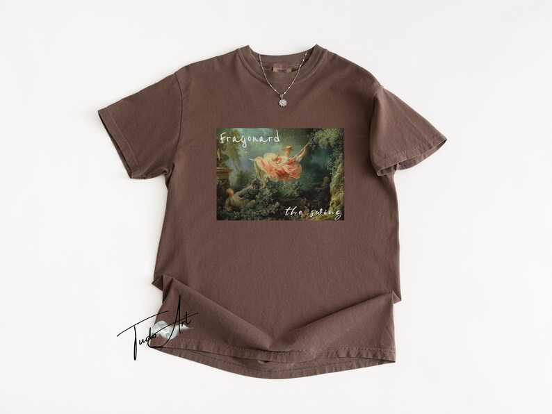 May include: A brown t-shirt featuring a print of a classical painting with the words "Fragonard" and "the swing". A silver necklace with a pendant is visible. The shirt is laid flat on a white surface.