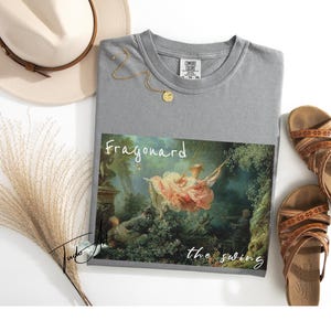 May include: Gray t-shirt featuring a print of Fragonard's "The Swing" painting. The shirt is styled with a beige hat, gold necklace, and brown sandals. The text "Fragonard the swing" is printed on the shirt.
