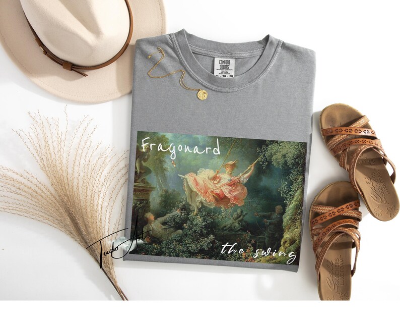 May include: Gray t-shirt featuring a print of Fragonard's "The Swing" painting. The shirt is styled with a beige hat, gold necklace, and brown sandals. The text "Fragonard the swing" is printed on the shirt.