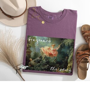 May include: A purple t-shirt featuring a print of Fragonard's "The Swing" painting. The shirt is accompanied by brown sandals, a beige hat, and a gold necklace. The text "Fragonard the swing" is printed on the shirt.