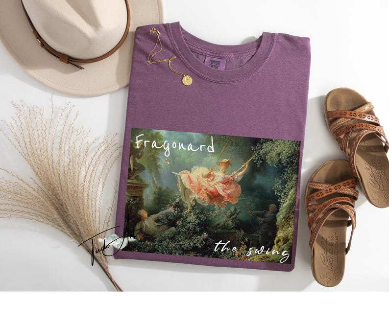 May include: A purple t-shirt featuring a print of Fragonard's "The Swing" painting. The shirt is accompanied by brown sandals, a beige hat, and a gold necklace. The text "Fragonard the swing" is printed on the shirt.
