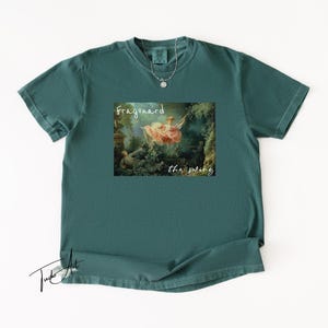 May include: Teal-colored t-shirt featuring a framed print of Fragonard's "The Swing" painting. The text "Fragonard" and "the swing" are printed below the image. A silver necklace with a small pendant is visible.