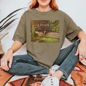 May include: An olive green t-shirt featuring the text "I'm literally fine" above a painting of a turtle in a pond. The model is wearing blue jeans and white trainers.