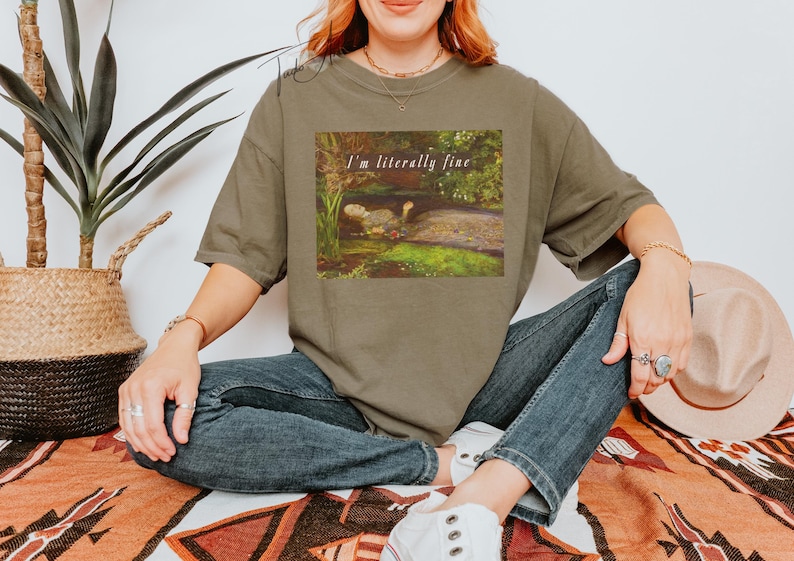 May include: An olive green t-shirt featuring the text "I'm literally fine" above a painting of a turtle in a pond. The model is wearing blue jeans and white trainers.