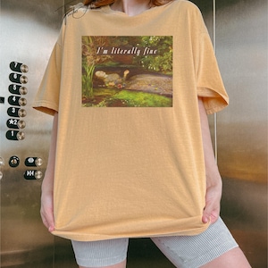 May include: Mustard yellow t-shirt with a rectangular graphic showing a painting of a person in water, surrounded by greenery. The text "I'm literally fine" is above the painting. The shirt has short sleeves and a relaxed fit.