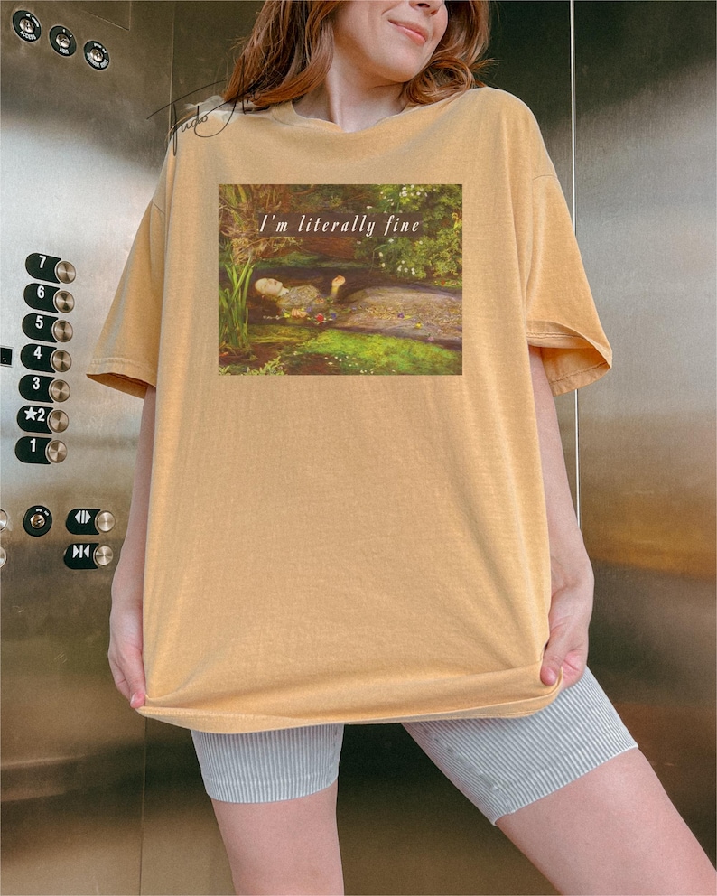 May include: Mustard yellow t-shirt with a rectangular graphic showing a painting of a person in water, surrounded by greenery. The text "I'm literally fine" is above the painting. The shirt has short sleeves and a relaxed fit.