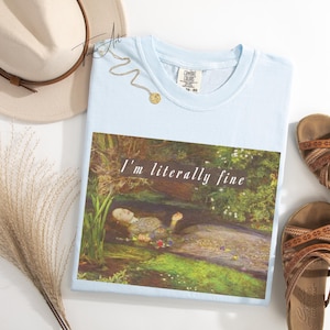 May include: Pale blue t-shirt featuring the text "I'm literally fine" above a painting of a woman in water. A beige hat, sandals, and a gold necklace are also visible in the image.