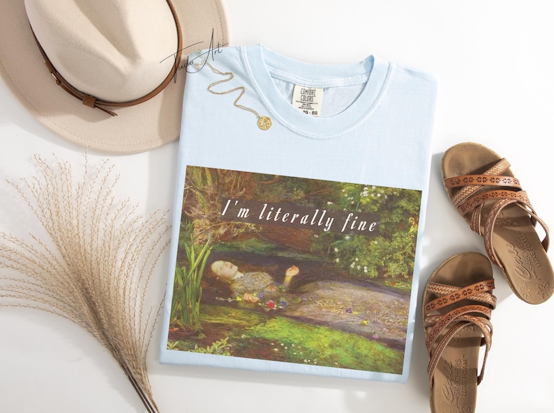 May include: Pale blue t-shirt featuring the text "I'm literally fine" above a painting of a woman in water. A beige hat, sandals, and a gold necklace are also visible in the image.