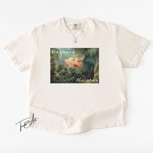 May include: Cream-colored t-shirt featuring a framed print of Fragonard's "The Swing" artwork. The print includes the text "Fragonard" above and "the swing" below the image. A silver necklace is visible.