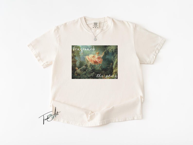 May include: Cream-colored t-shirt featuring a framed print of Fragonard's "The Swing" artwork. The print includes the text "Fragonard" above and "the swing" below the image. A silver necklace is visible.