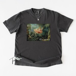 May include: Dark gray t-shirt featuring a print of Fragonard's "The Swing" painting. The artwork depicts a woman on a swing in a garden setting. The text "Fragonard the swing" is printed above and below the image in a white script font.