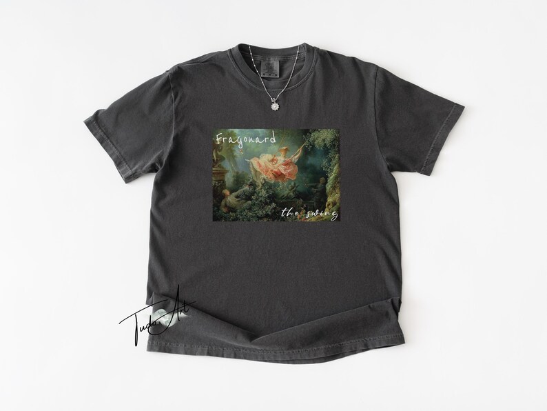 May include: Dark gray t-shirt featuring a print of Fragonard's "The Swing" painting. The artwork depicts a woman on a swing in a garden setting. The text "Fragonard the swing" is printed above and below the image in a white script font.