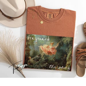 May include: Rust-colored t-shirt featuring a print of Fragonard's "The Swing" painting. The shirt is paired with brown sandals, a beige hat, and a gold necklace. The text "Fragonard the swing" is printed on the shirt.