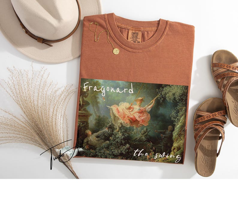 May include: Rust-colored t-shirt featuring a print of Fragonard's "The Swing" painting. The shirt is paired with brown sandals, a beige hat, and a gold necklace. The text "Fragonard the swing" is printed on the shirt.