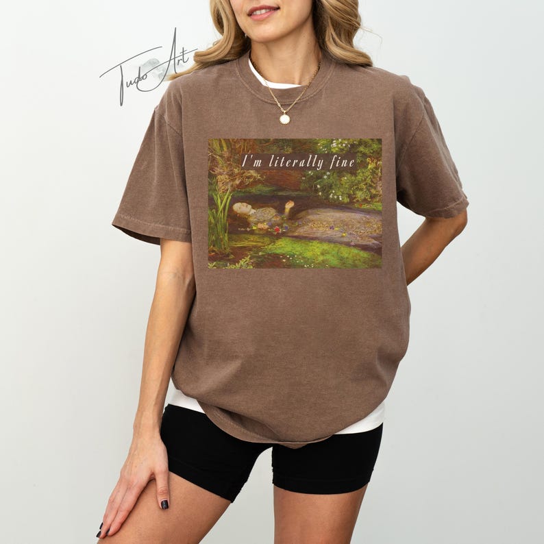 May include: Brown t-shirt with a white undershirt and black shorts. The t-shirt features a framed image of a painting with the text "I'm literally fine". The painting depicts a person in a pond.