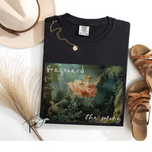 May include: Black t-shirt featuring a print of Fragonard's "The Swing" painting. The shirt is styled with a gold necklace, brown sandals, and a beige hat. A bundle of dried ornamental grass is also included.