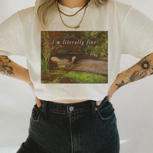 May include: Cream-coloured t-shirt with a square graphic of a painting depicting a person in a pond. The text "I'm literally fine" is above the painting. The person is wearing black jeans.