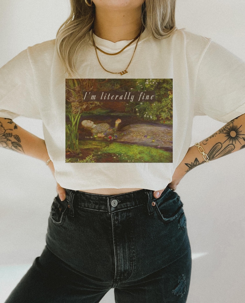 May include: Cream-coloured t-shirt with a square graphic of a painting depicting a person in a pond. The text "I'm literally fine" is above the painting. The person is wearing black jeans.