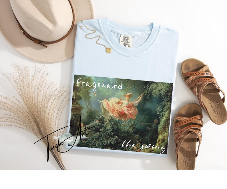 May include: Light blue t-shirt featuring the painting "The Swing" by Fragonard. The shirt has a crew neck and short sleeves. A gold necklace and brown sandals are also in the image, along with a beige hat and decorative grass.