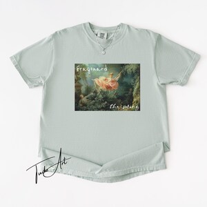 May include: A light green t-shirt featuring a framed print of a classical painting, with the words "Fragonard" and "the swing" in white script. The shirt has a crew neck and short sleeves.