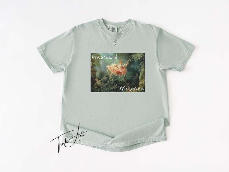 May include: A light green t-shirt featuring a framed print of a classical painting, with the words "Fragonard" and "the swing" in white script. The shirt has a crew neck and short sleeves.