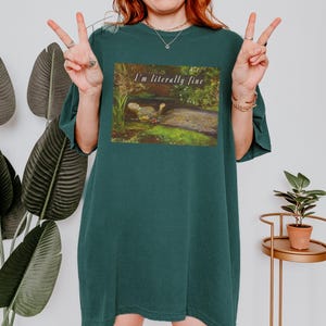 May include: Dark teal t-shirt with a rectangular graphic featuring a painting of a dodo bird in a natural setting. The text "I'm literally fine" is above the painting. The shirt is a relaxed fit.