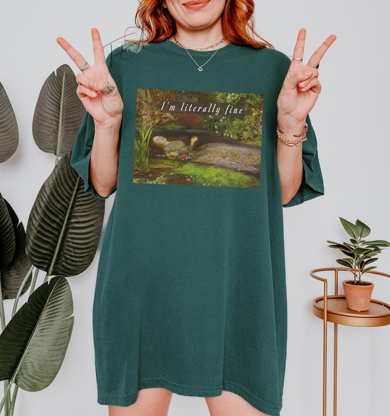 May include: Dark teal t-shirt with a rectangular graphic featuring a painting of a dodo bird in a natural setting. The text "I'm literally fine" is above the painting. The shirt is a relaxed fit.