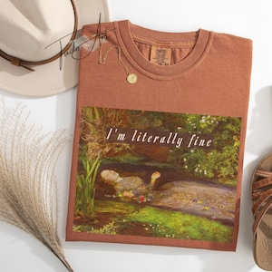 May include: A terracotta-coloured t-shirt with the words "I'm literally fine" printed on the front, featuring a painting of a woman in water. A beige hat, sandals, and a gold necklace are also visible.