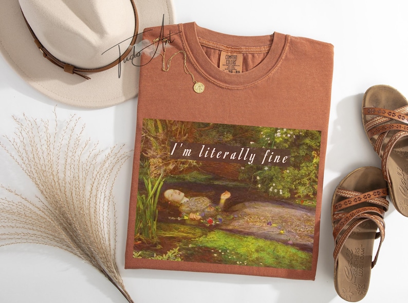 May include: A terracotta-coloured t-shirt with the words "I'm literally fine" printed on the front, featuring a painting of a woman in water. A beige hat, sandals, and a gold necklace are also visible.