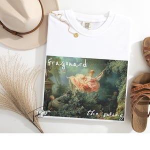 May include: White t-shirt featuring a print of Fragonard's "The Swing" painting. The shirt is paired with brown sandals, a beige hat, and a gold necklace. The artwork depicts a woman on a swing in a garden setting.